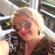 Blonde female wearing sunglasses and a big warm smile and bright red lipstick. She is taking a selfie and is wearing an gem embossed top.