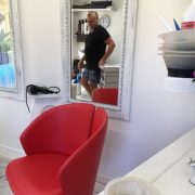 Male hairdresser wearing a black top and jean shorts. Featured is a big red chair and a mirror where you can see his reflection.